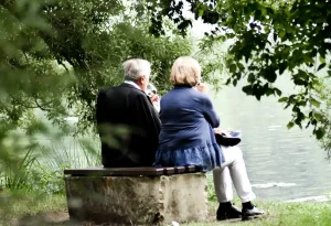 Two elderly people sitting on a bench near a calm lake, surrounded by green trees in Bedford, NH 